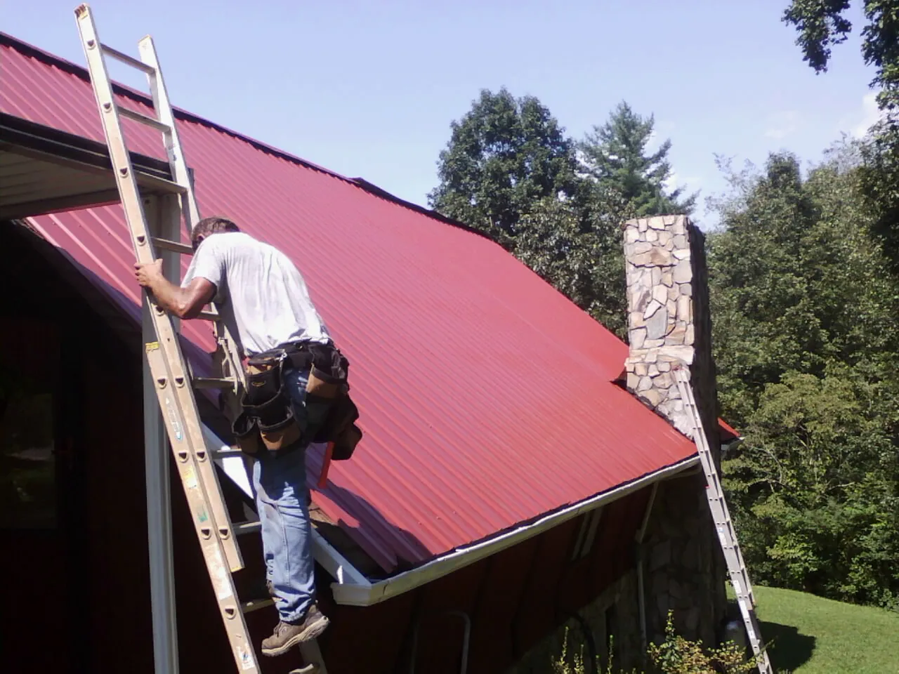 Roofer on a ladder beside a red metal roof and stone chimney — Soffit & Fascia Repair by Roofing Brothers in Fort Monmouth
